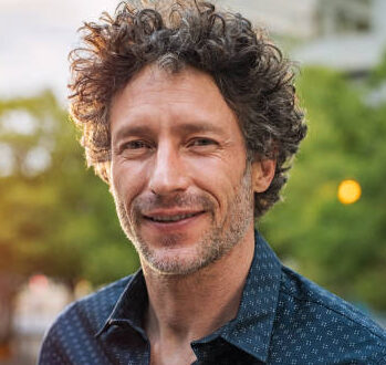 smiling man with dark curly hair outdoors.